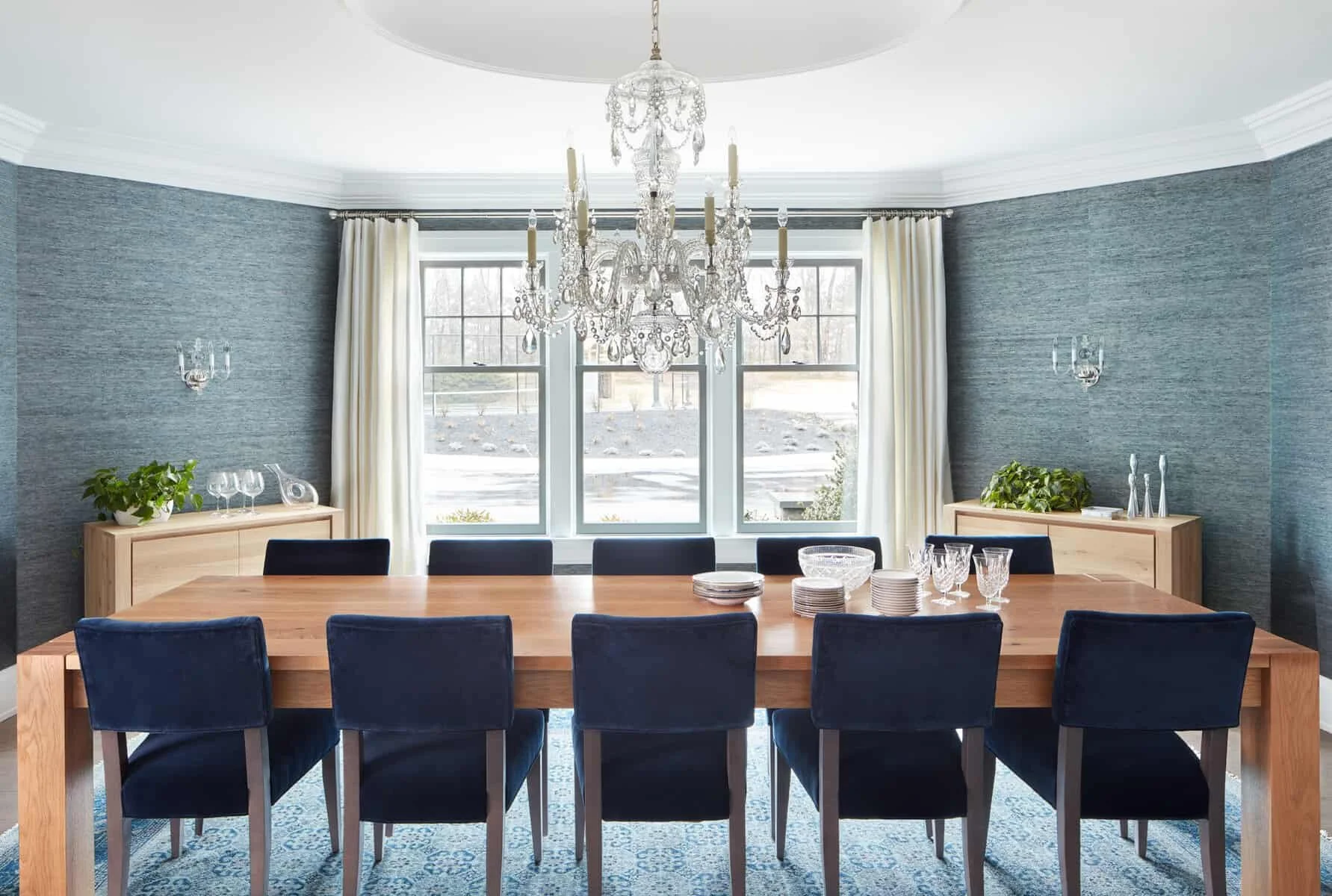 A modern dining room with a large wooden table set with glassware and plates, surrounded by blue upholstered chairs, featuring a chandelier hanging from the ceiling, large windows with white curtains, two light wood sideboards with plants and decorat