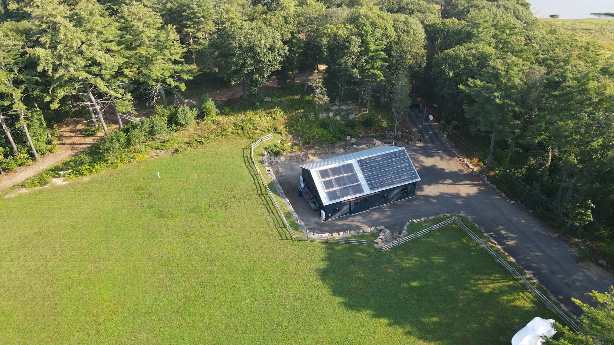An aerial view of a small dark-colored building with solar panels on its roof, surrounded by green grass, trees, a gravel pathway, and a white fence, in a rural setting.