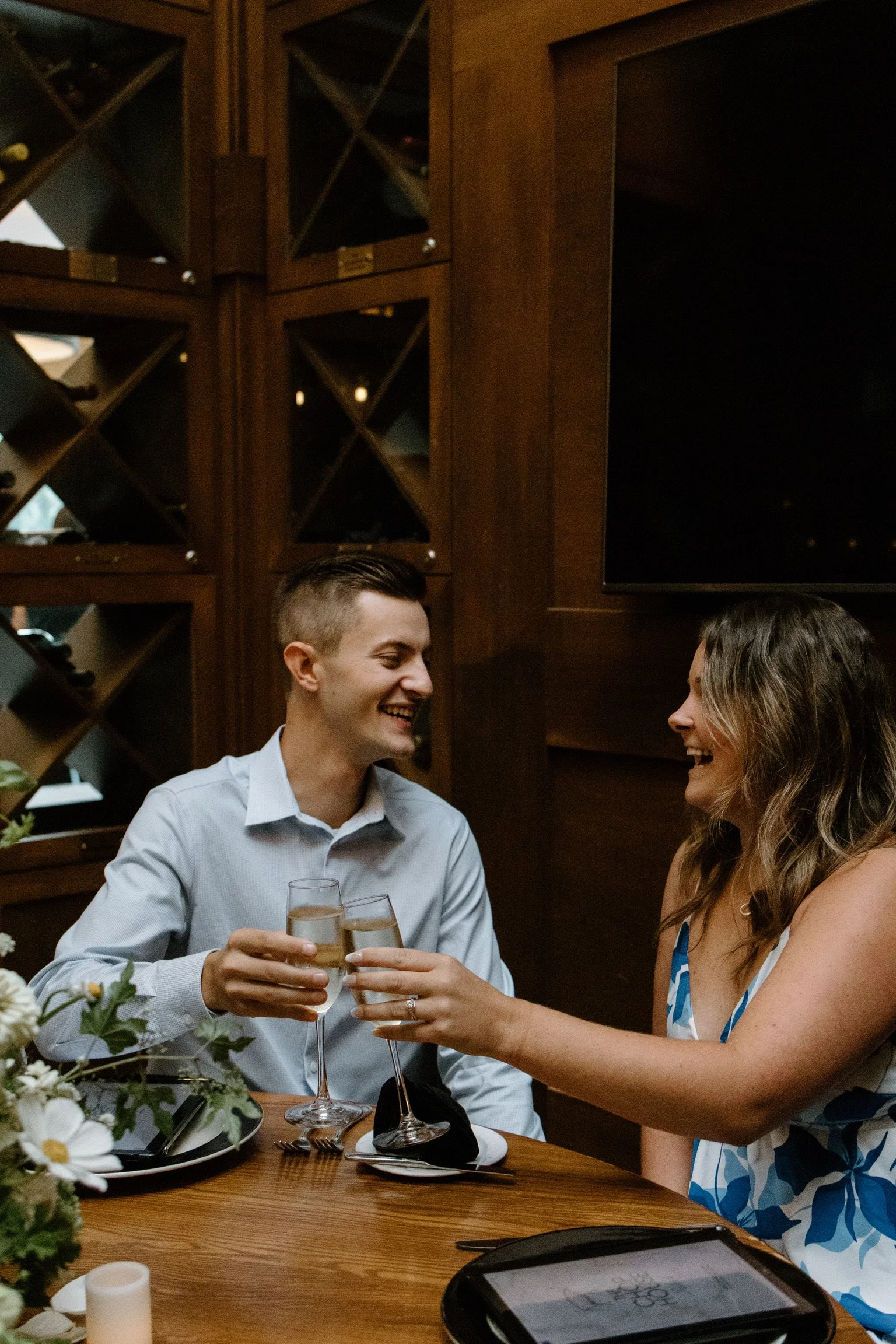 A man and woman sharing a toast with champagne at a dining table, smiling and laughing, in a warmly lit restaurant with wooden decor and wine shelves in the background.