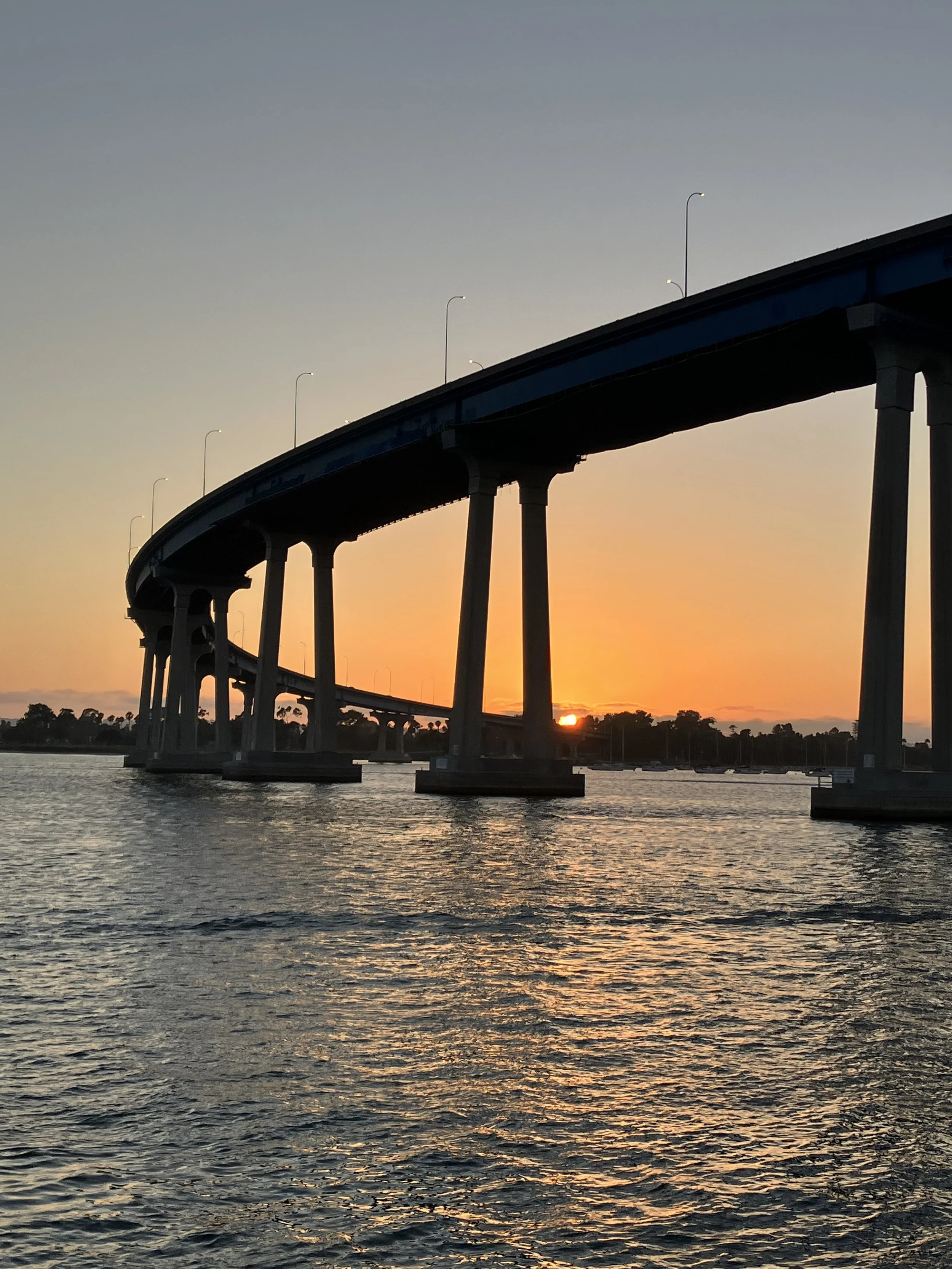 A bridge over water at sunset with the sun near the horizon and trees in the background.
