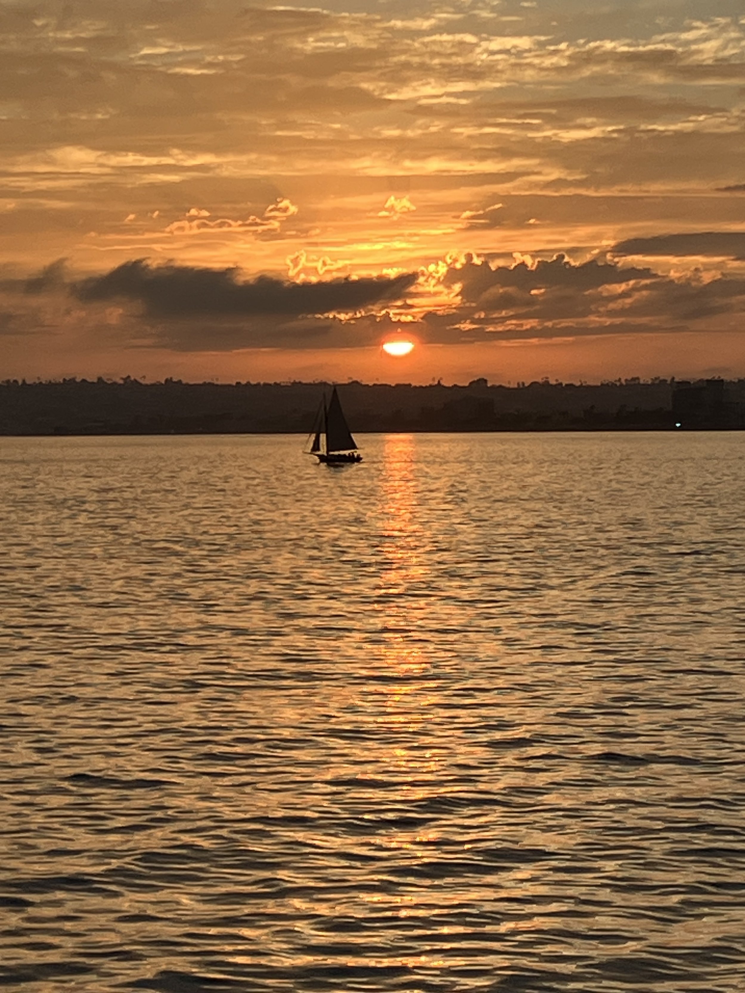 Sunset over the water with a sailboat, partly cloudy sky, and reflections on the surface.