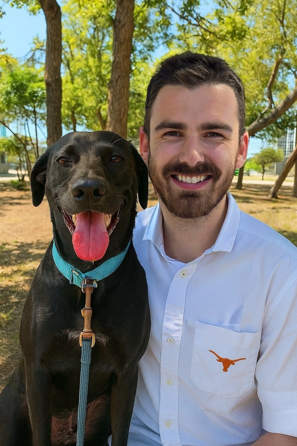 A smiling man in a white shirt sitting next to a happy black dog with a blue collar outside in a park with trees and a clear blue sky.
