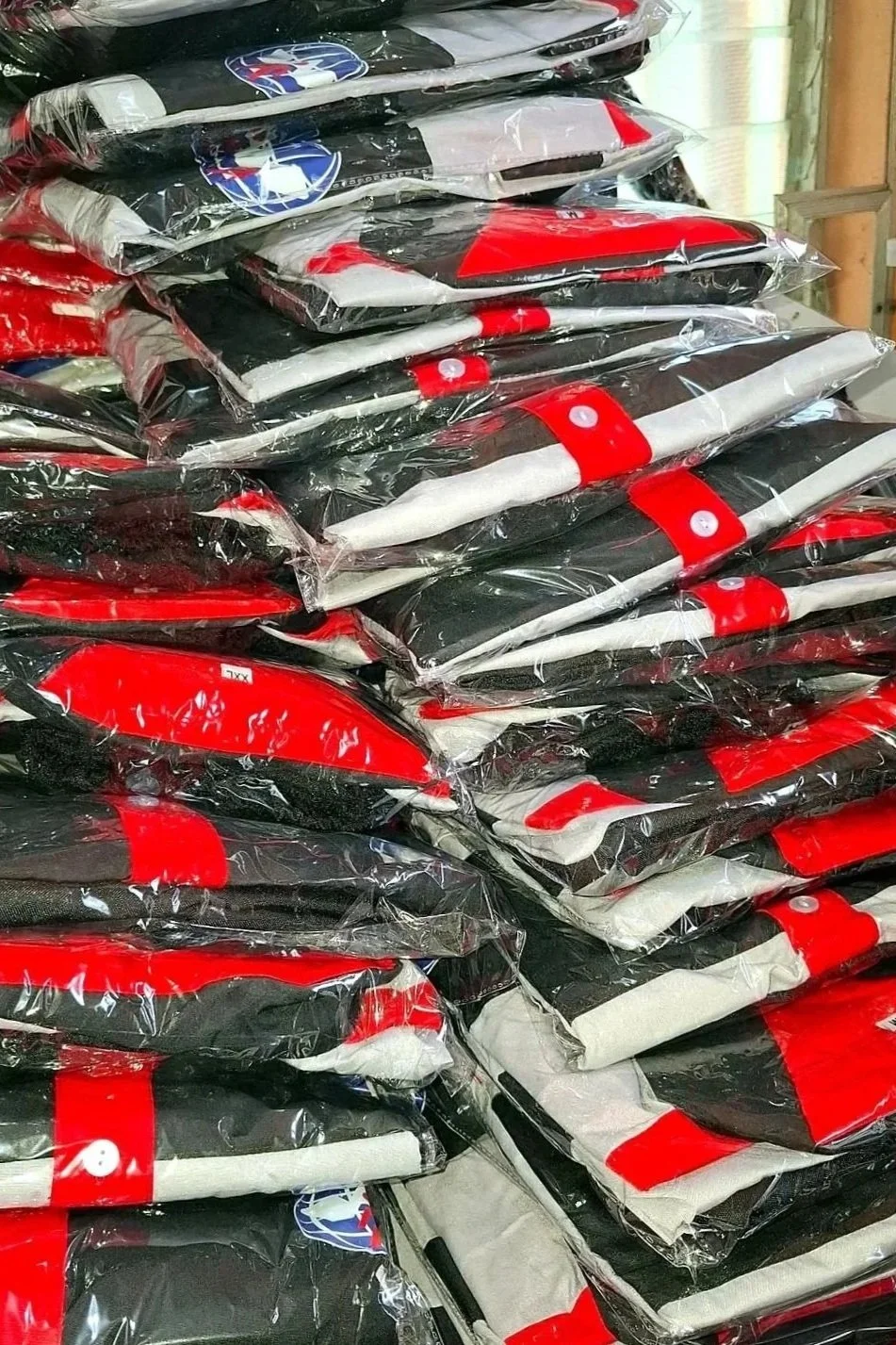 Stack of folded clothing or fabric items in plastic packaging, featuring red, black, and white colors, with some items displaying logos or markings.