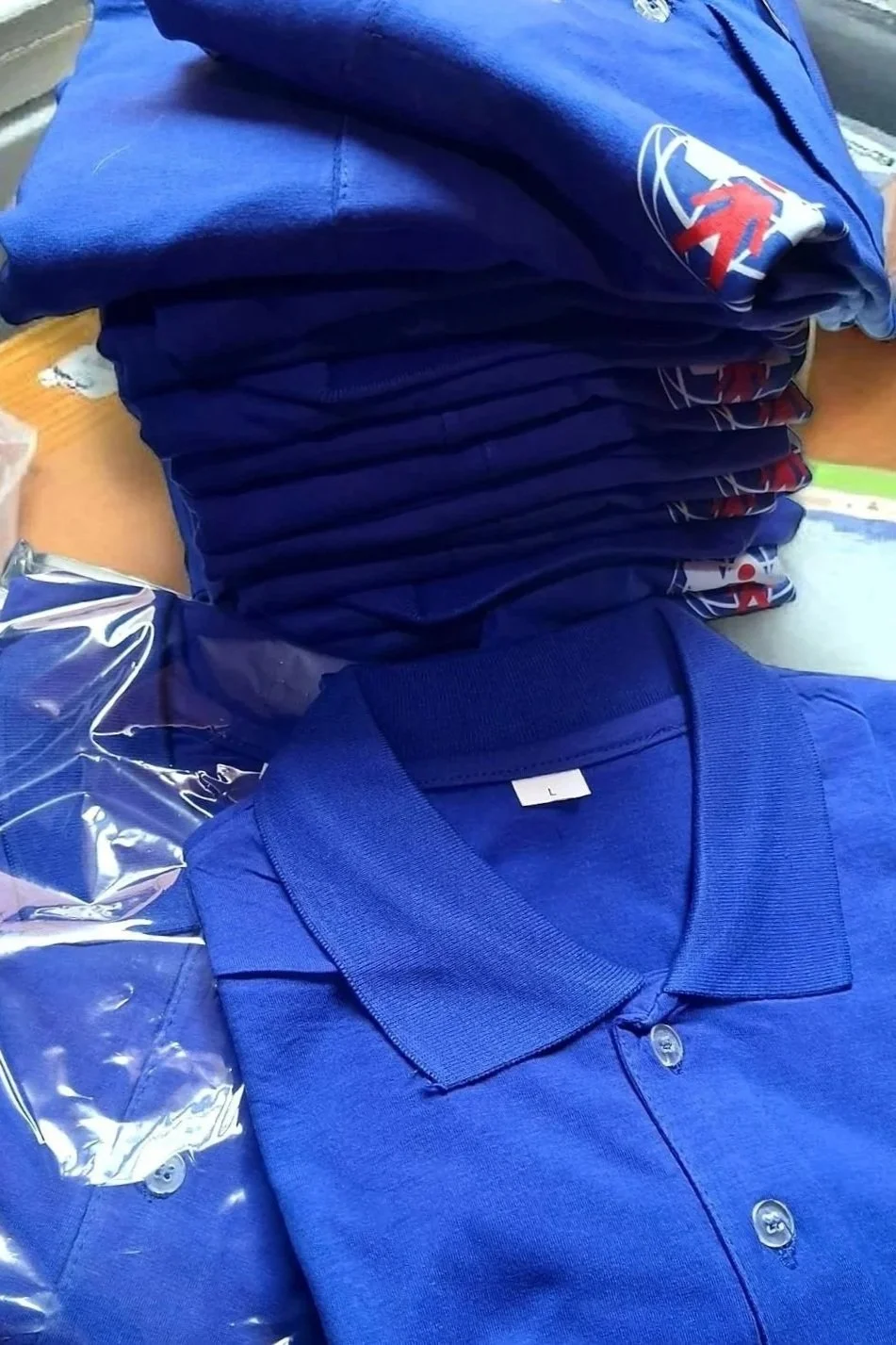 A stack of folded blue sports jerseys with team logos, and a folded blue polo shirt with a size 'L' tag on a wooden surface.