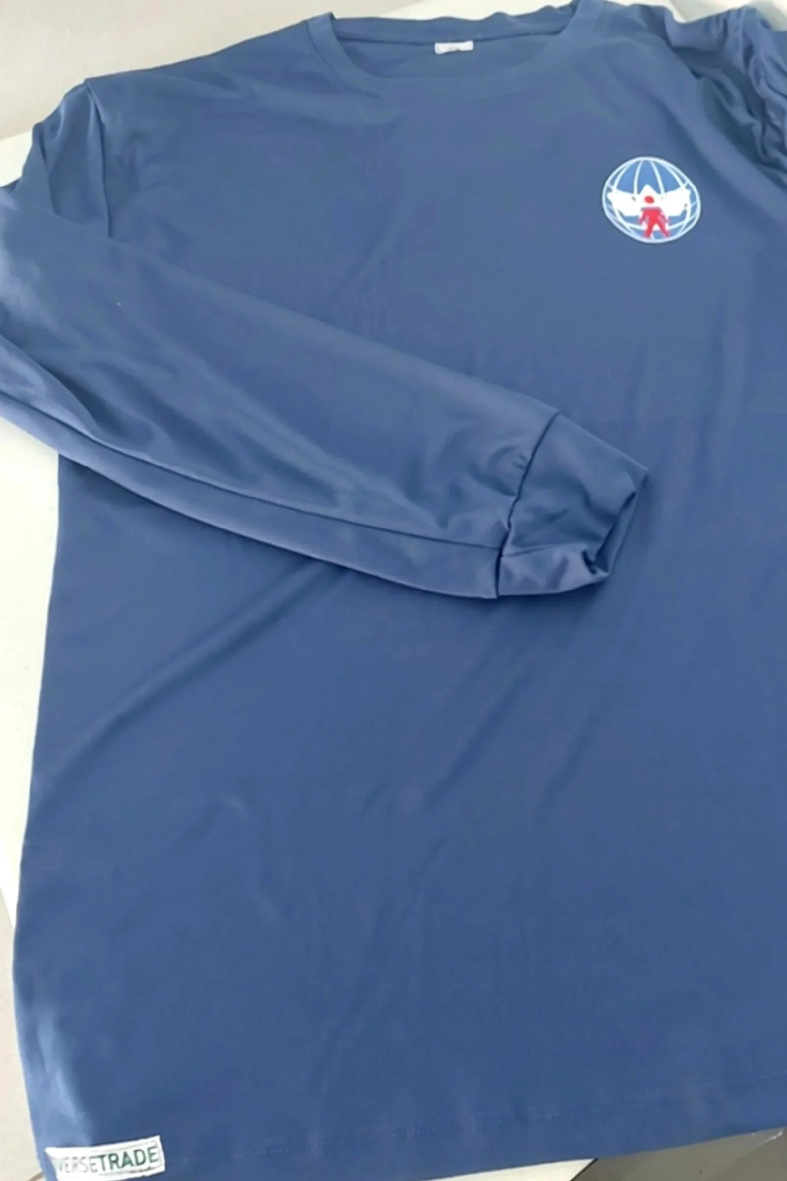 A blue long-sleeve shirt with a logo on the upper left chest area featuring a globe with a red figure and a white dove, lying on a flat surface, with a small white and green label at the bottom left corner reading 'DIVERSETRADE'.