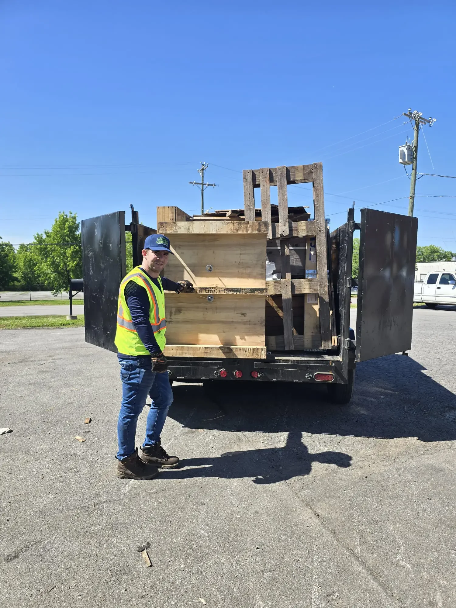 A smiling man wearing a yellow safety vest, black gloves, and a blue cap stands next to an open truck filled with wooden pallets and containers, under a clear blue sky.