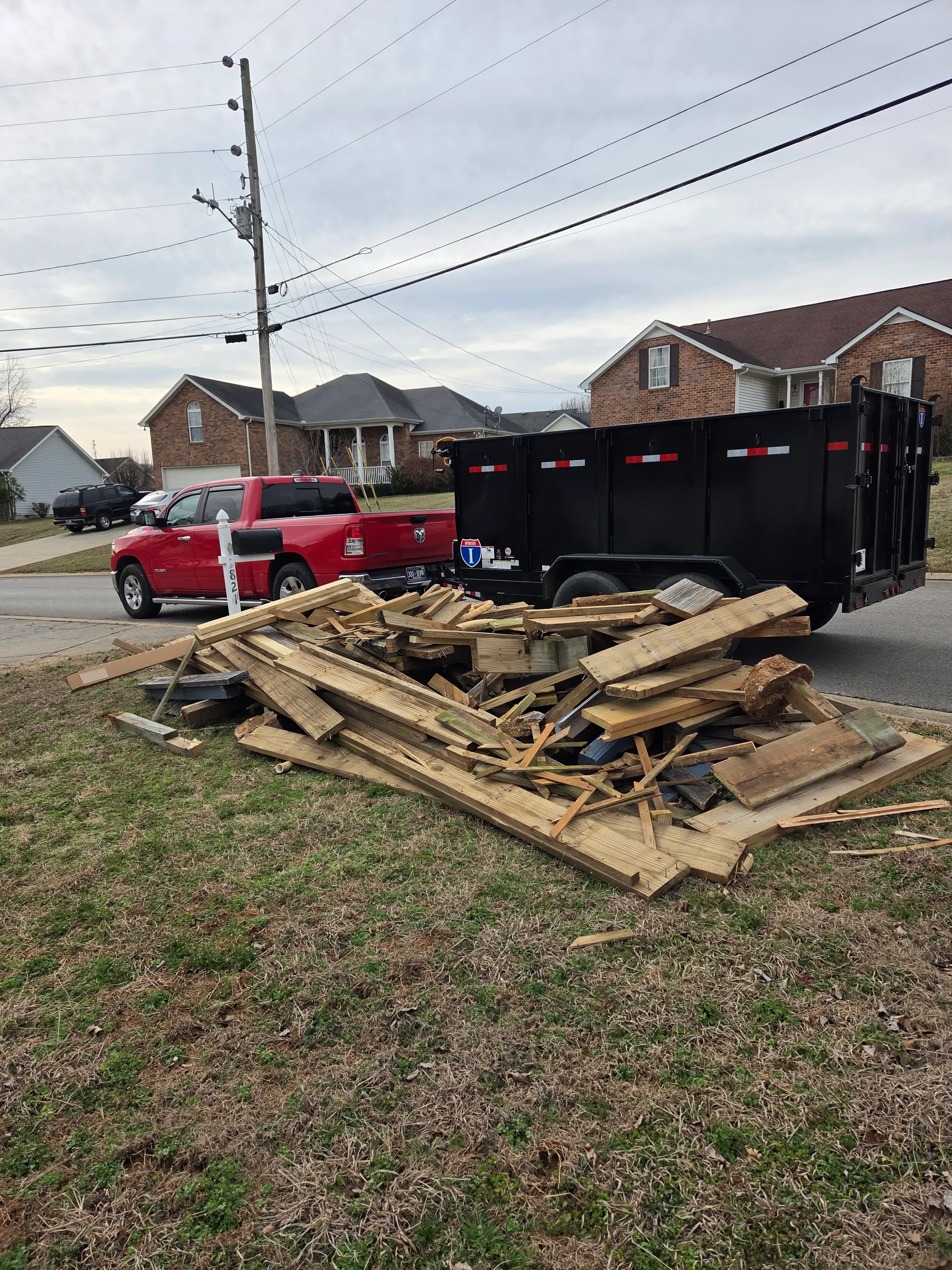 Pile of discarded wooden boards and debris on a grassy area near a suburban street, with houses and a utility pole in the background.