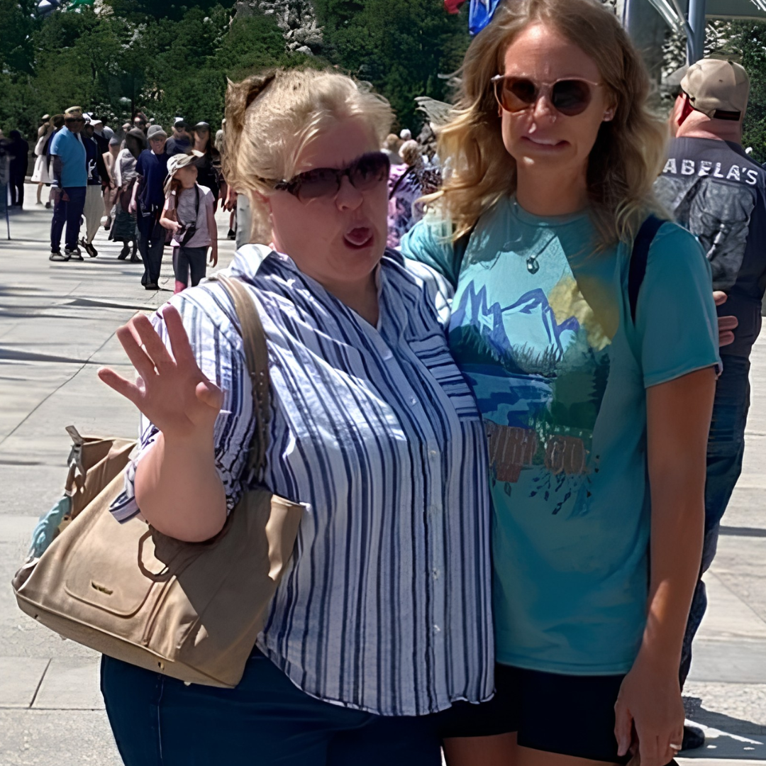 Two women with sunglasses posing outdoors in a crowded area, with trees and people in the background.