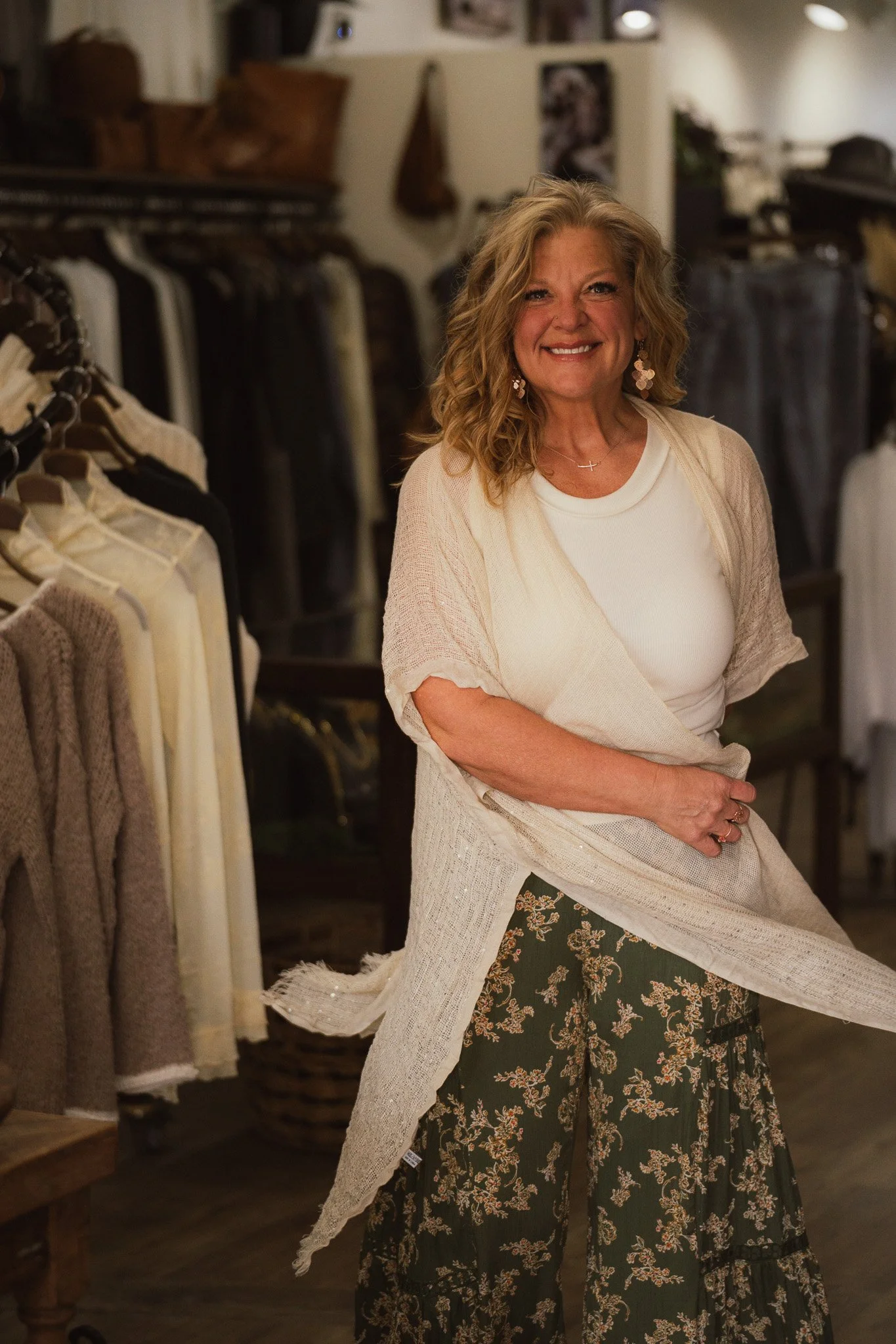 A smiling woman with blonde curly hair wearing a white top and floral wide-leg pants, standing in a clothing store surrounded by racks of sweaters and garments.