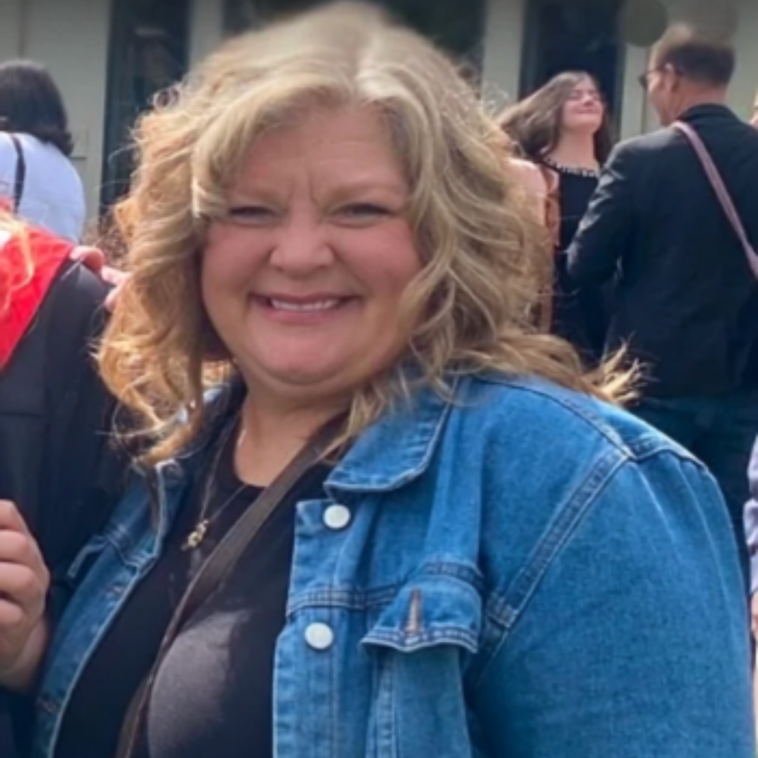 Smiling woman with blonde curly hair wearing a denim jacket, standing among a crowd of people outdoors.