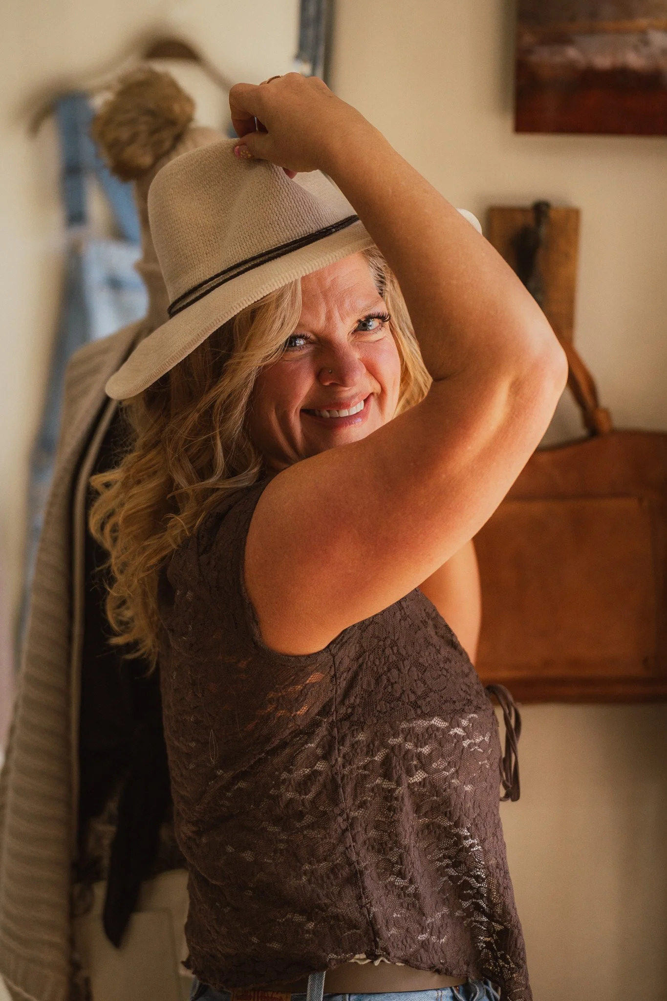 A woman with blonde curly hair lifting her arm, smiling, and wearing a beige hat and a dark lace top. She stands indoors near a mirror and wooden furniture.