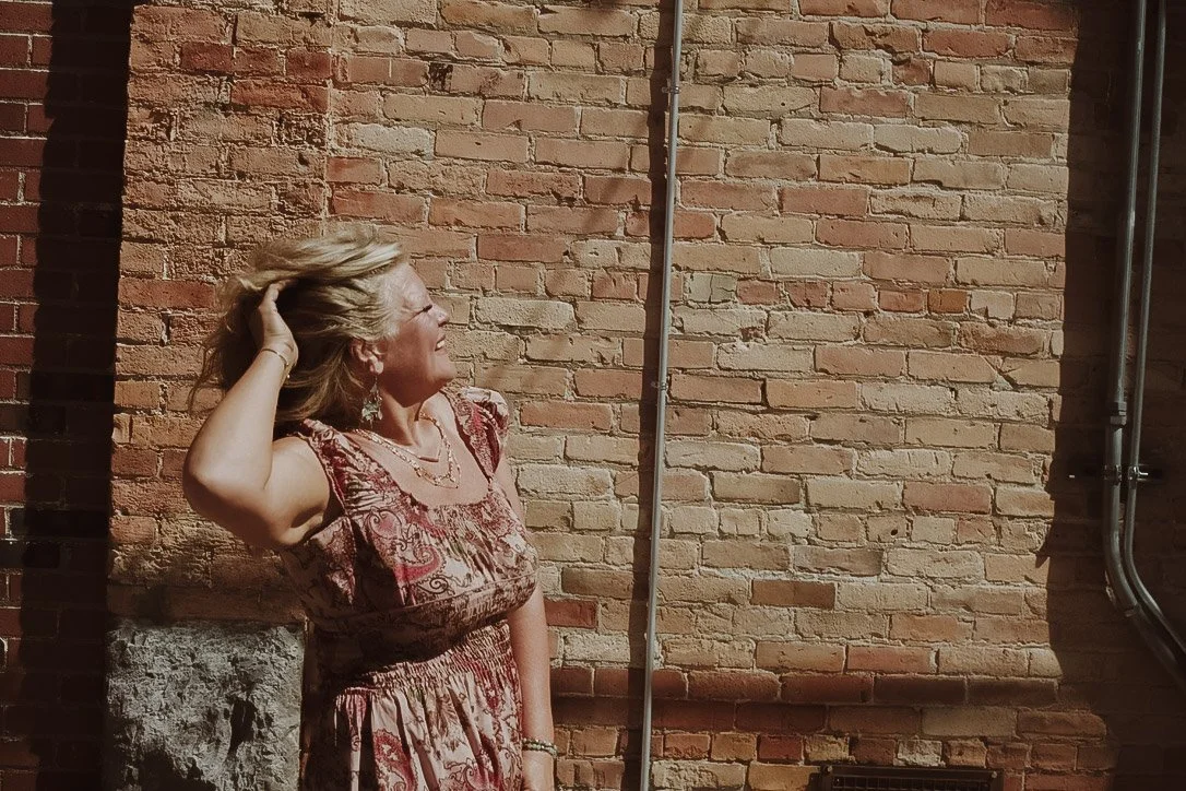 A woman with blonde hair and earrings standing against a brick wall, smiling and tilting her head back with her hand in her hair.