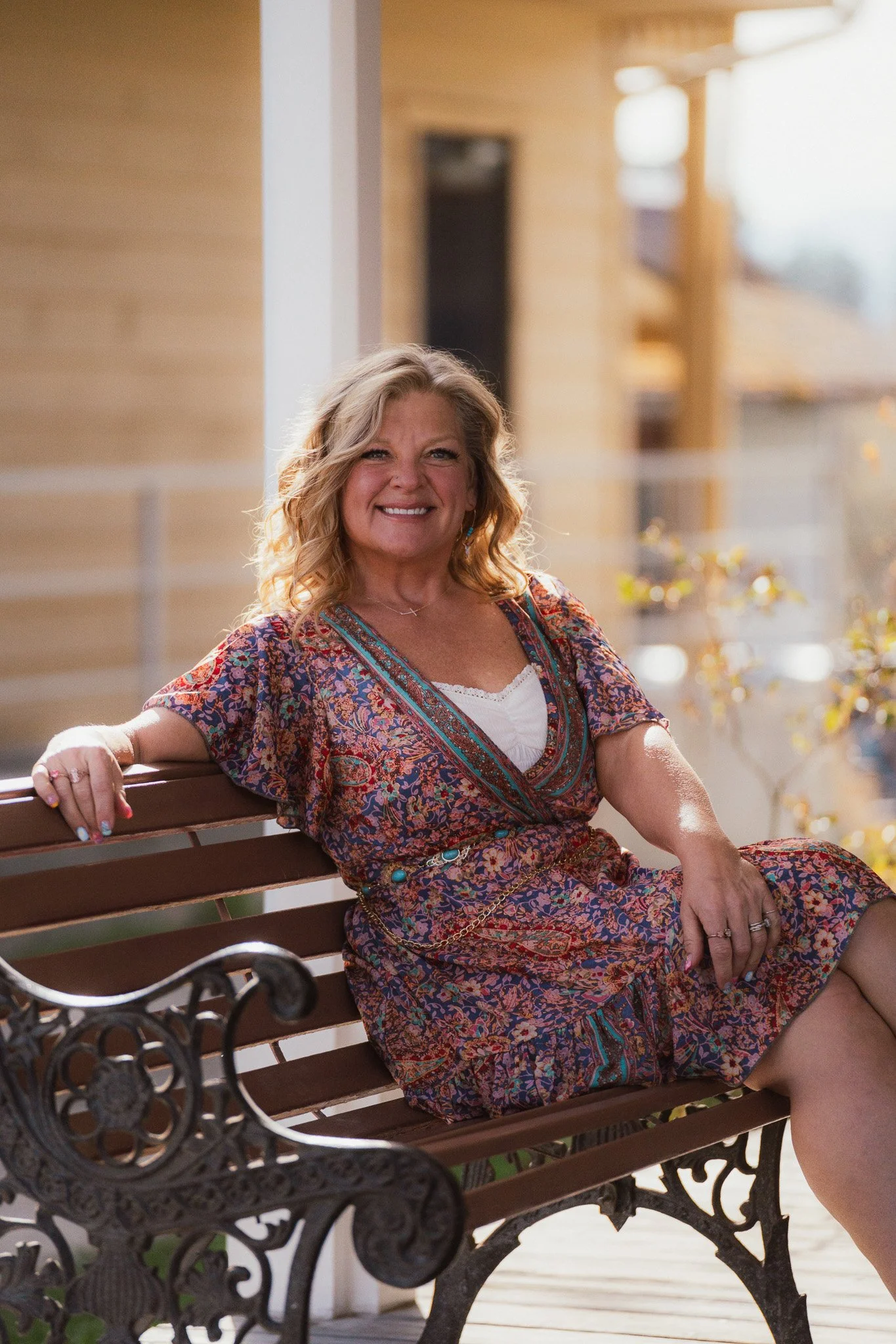 A woman with wavy blonde hair, smiling, sitting on a wooden park bench next to a black ornate wrought iron armrest, wearing a colorful patterned dress, outdoors in a sunny environment.