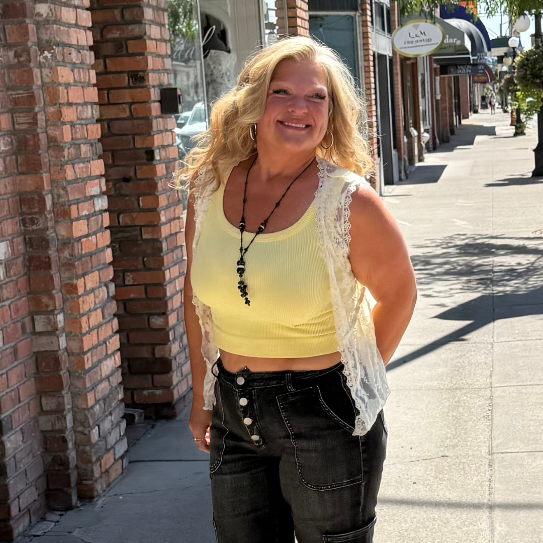 A woman with blonde hair wearing a yellow crop top, lace vest, black jeans, and beaded jewelry standing on a sunny city sidewalk smiling.