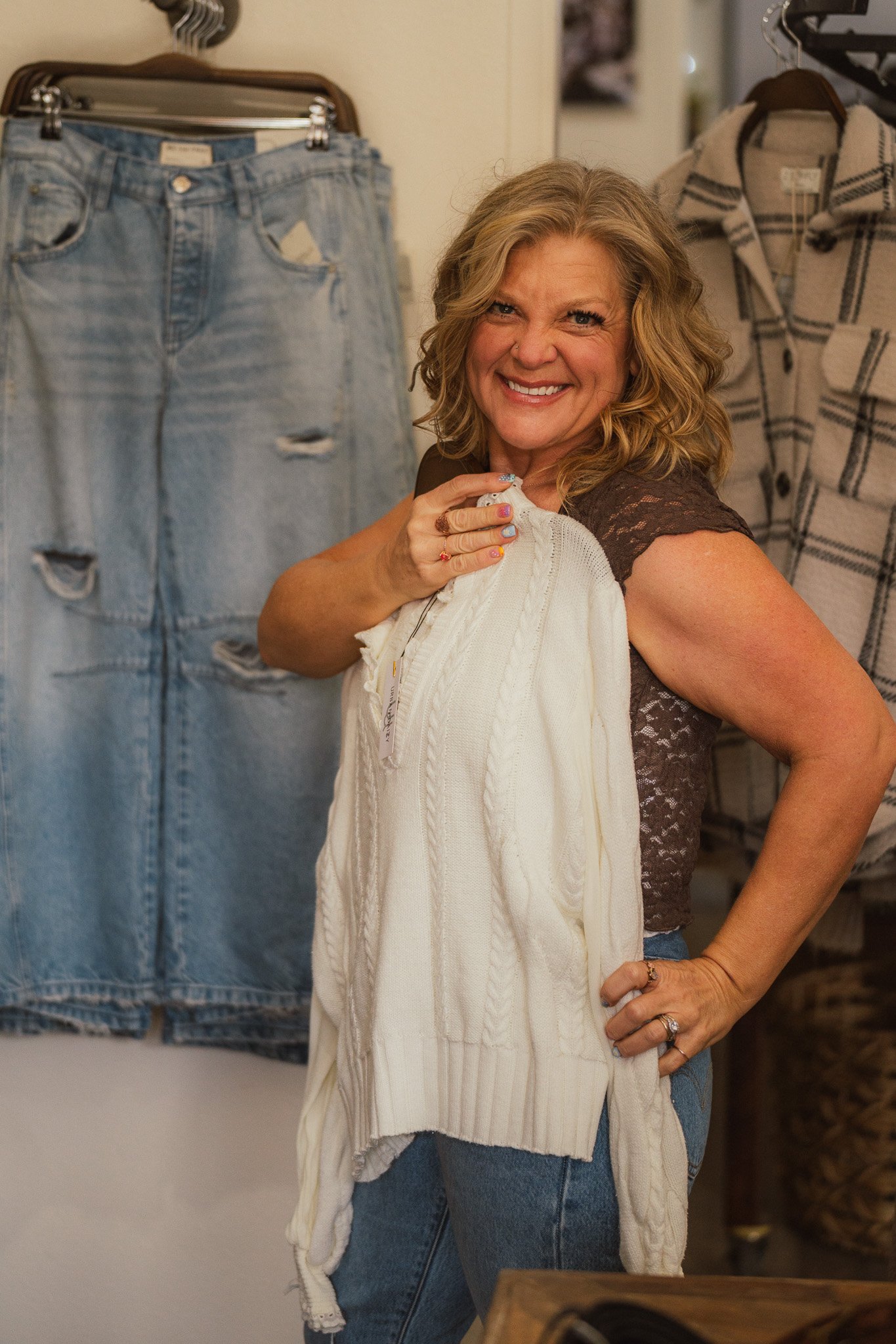 A smiling woman with curly blonde hair trying on a white knit sweater in a clothing store. Denim jeans and hanging clothes are visible in the background.