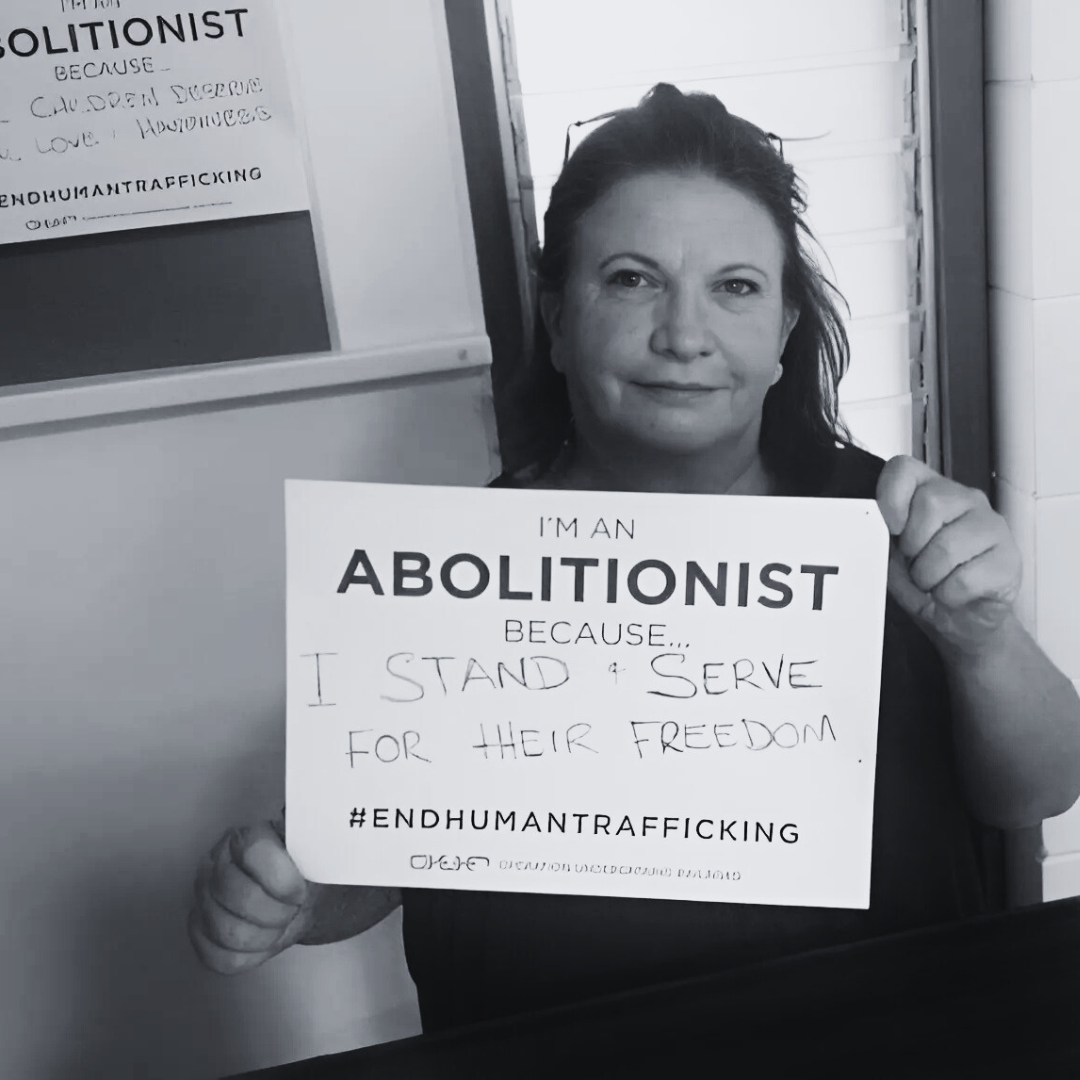 A woman holding a sign that reads, 'I'm an abolitionist because I stand and serve for their freedom,' in a room with posters on the wall, promoting the end of human trafficking.