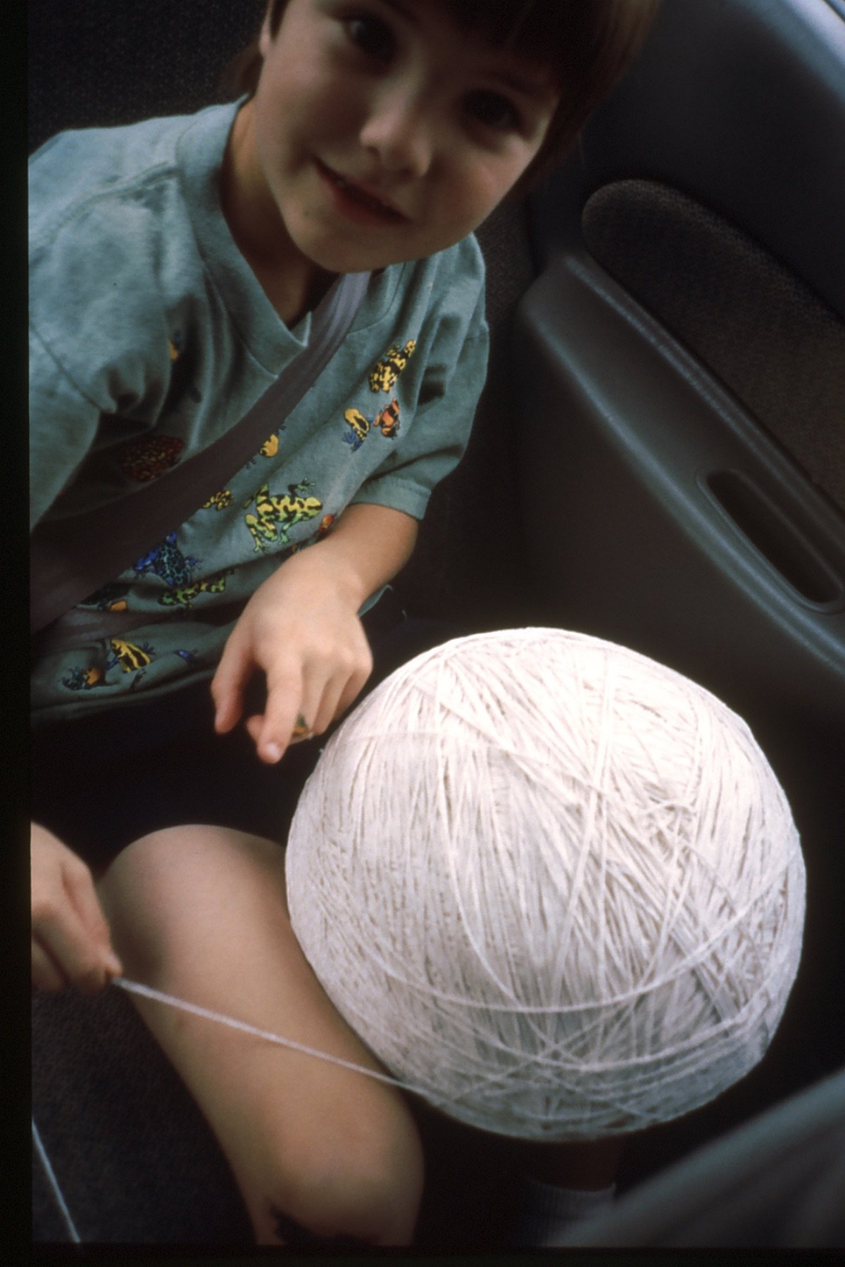 A young boy sitting in a car with a large white string ball on his lap, looking at the camera with a smile.