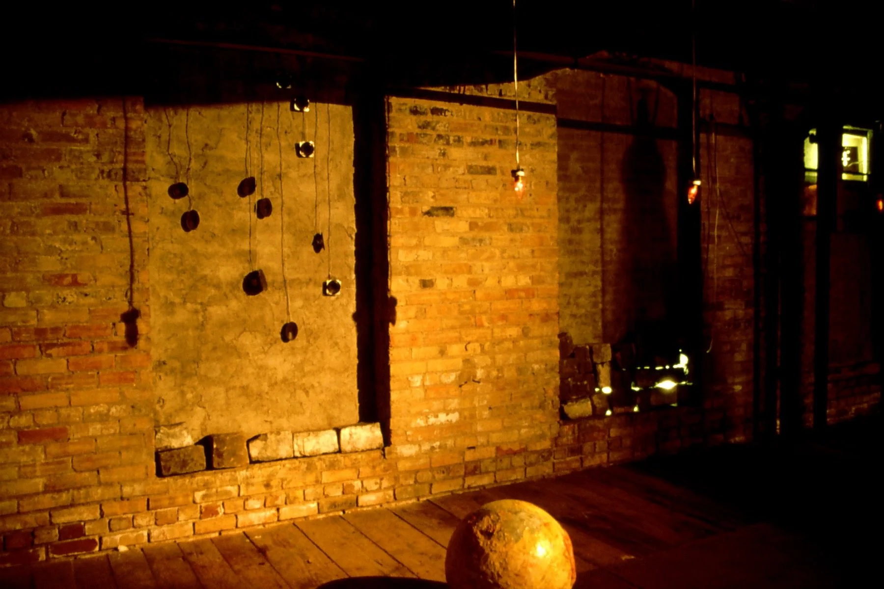 Dimly lit room with exposed brick walls, hanging lights, and a rusty spherical object on the wooden floor.