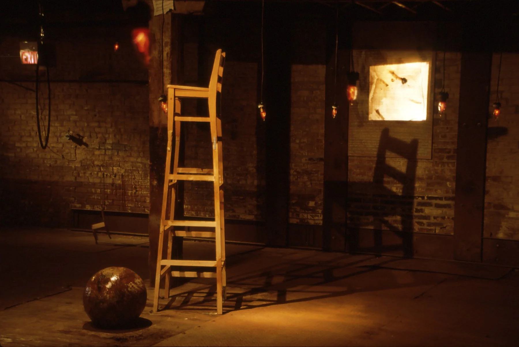 Interior scene with a wooden ladder, a rusted metal ball on the floor, brick walls, hanging light fixtures, and an illuminated framed picture or mirror on the wall. Shadows cast by the ladder and lights are visible.