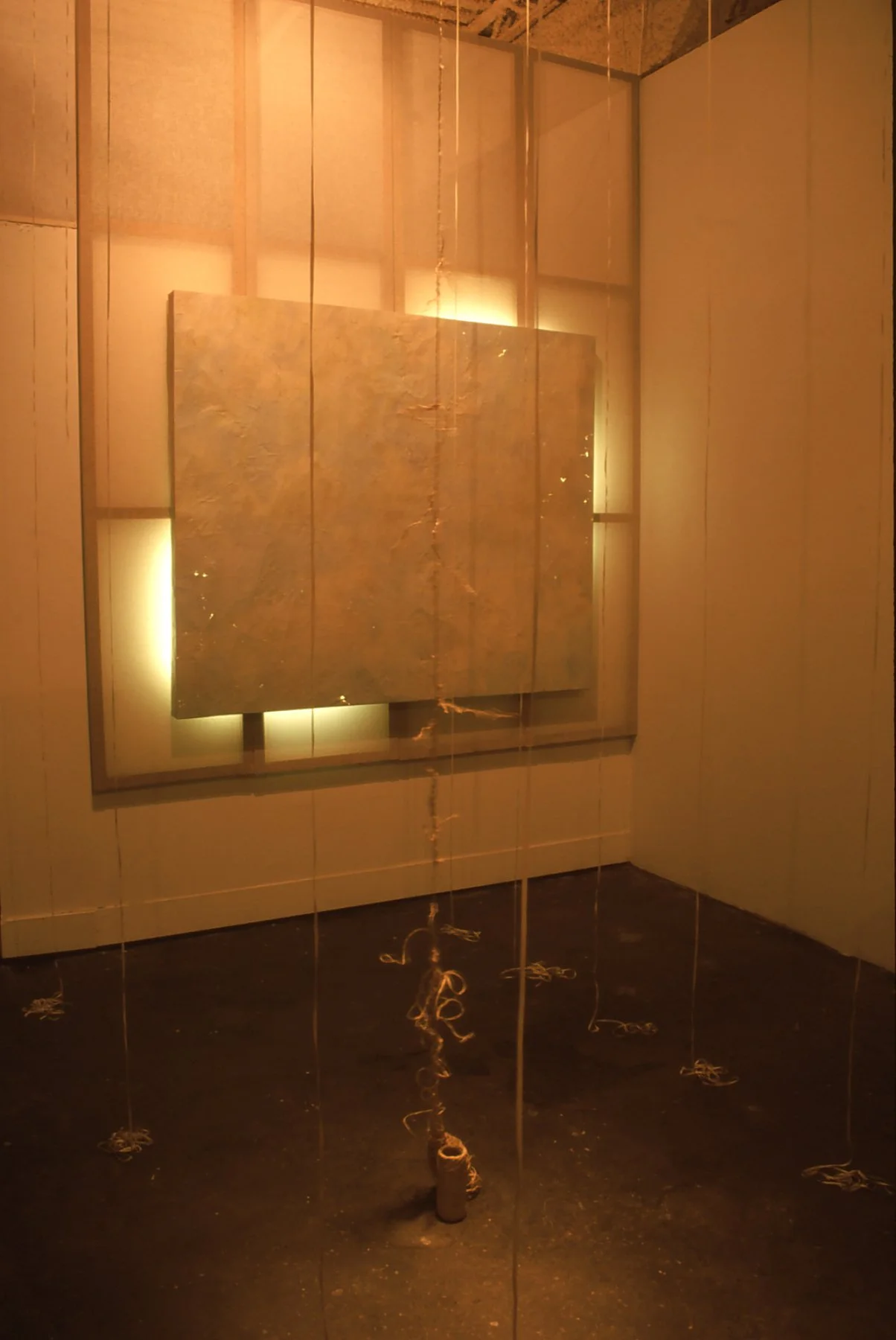 An interior art installation featuring a large rectangular stone or concrete slab leaning against a wall, illuminated with soft yellow lighting, with hanging strings or wires with paper snippets or paper-like objects dangling from the ceiling to the 