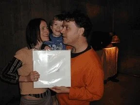 A woman holding a framed picture and a young boy are in conversation with a man, in an indoor setting at night.