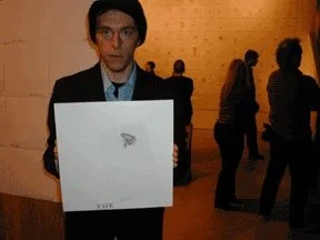 Person holding a white box with a small gray paper airplane logo, in an indoor setting with other people in the background.