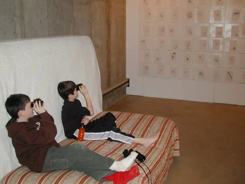 Two young boys sitting on a bed with vintage striped bedding, looking through binoculars, in a room with wooden walls and a display of small framed items or artwork.