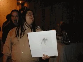 Two people at a party, one holding a drawing of a bird, the other making a surprised face in the background.