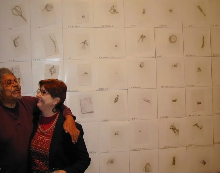 Two people are standing in front of a large chart on the wall with various objects attached, such as keys and small tools. They are smiling at each other, with one person's arm around the other's shoulder.