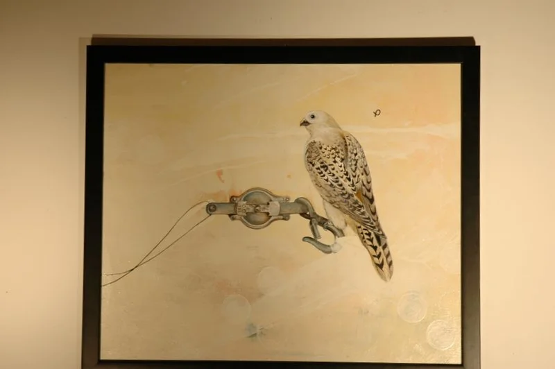 A framed painting or photo of a bird perched on a mechanical or metallic device against a light background.