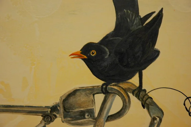 Painting of a black bird with yellow eyes and orange beak perched on a metallic bike handlebar against a yellow wall.