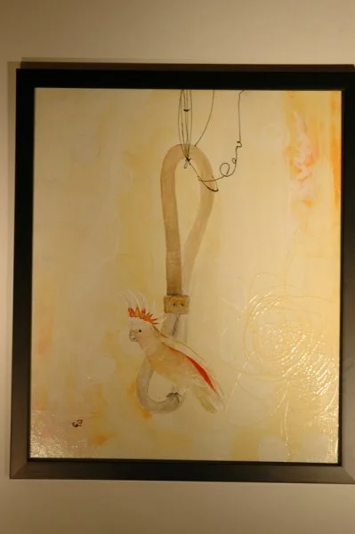 A framed abstract painting featuring a fish with orange and cream colors and a hook with a fishing line above it.