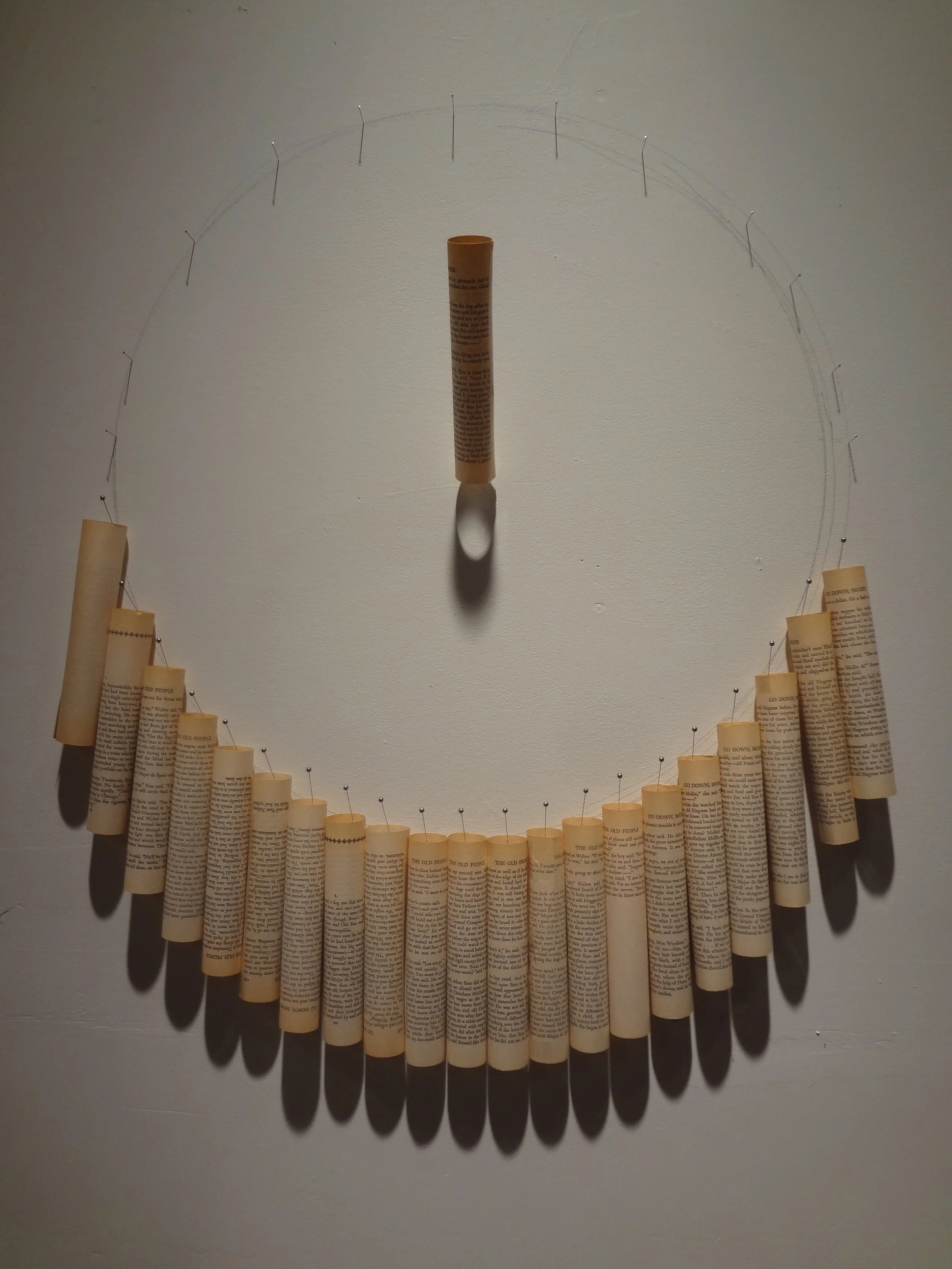 An art piece resembling a smiley face made with a large, circular wire and rolled-up pages pinned to it, with a vertical rolled-up page serving as the nose and a shadow cast underneath.