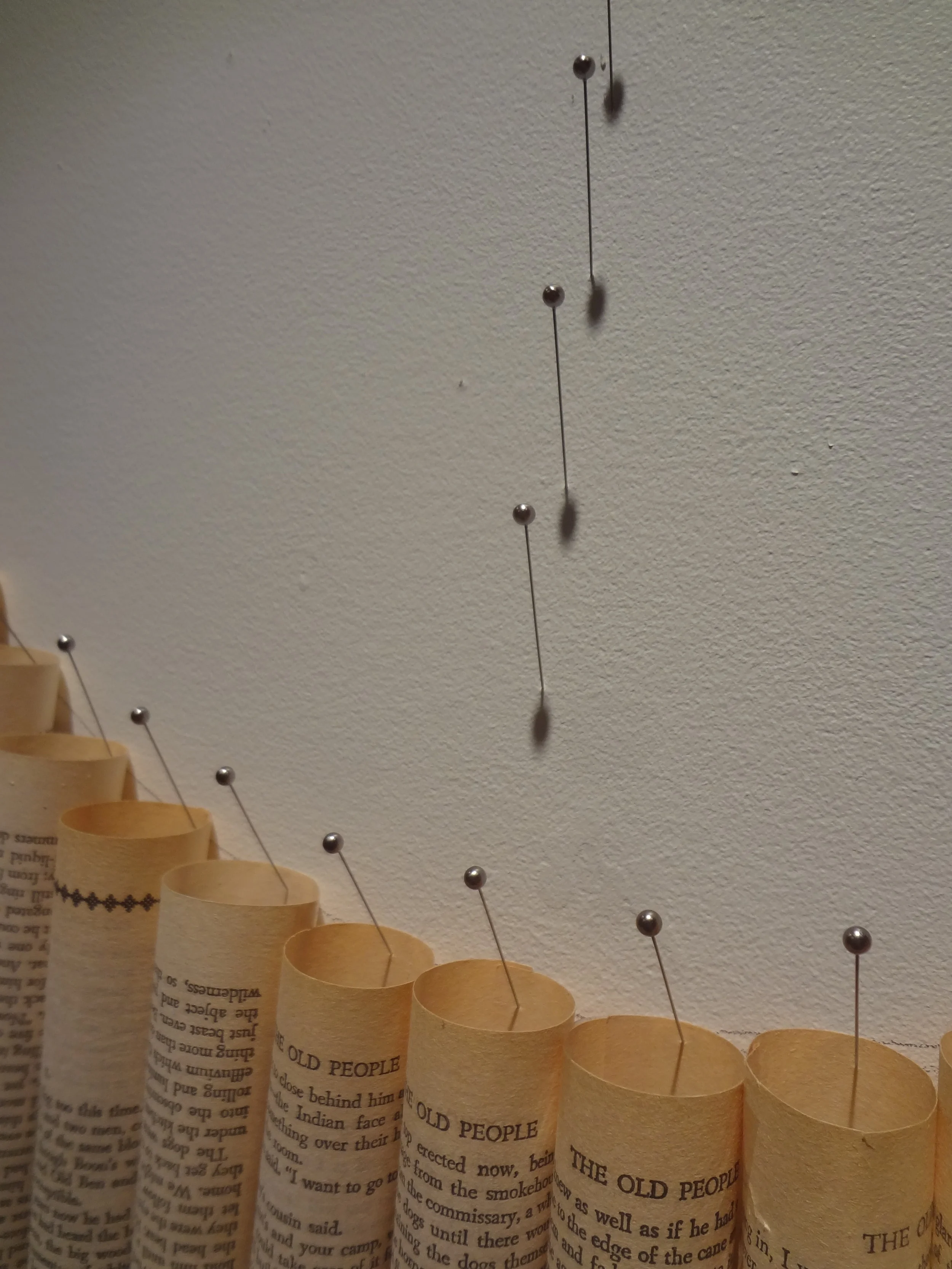 A paper newsprint scroll with pins inserted into the top, hanging from a ceiling, with a white wall in the background.