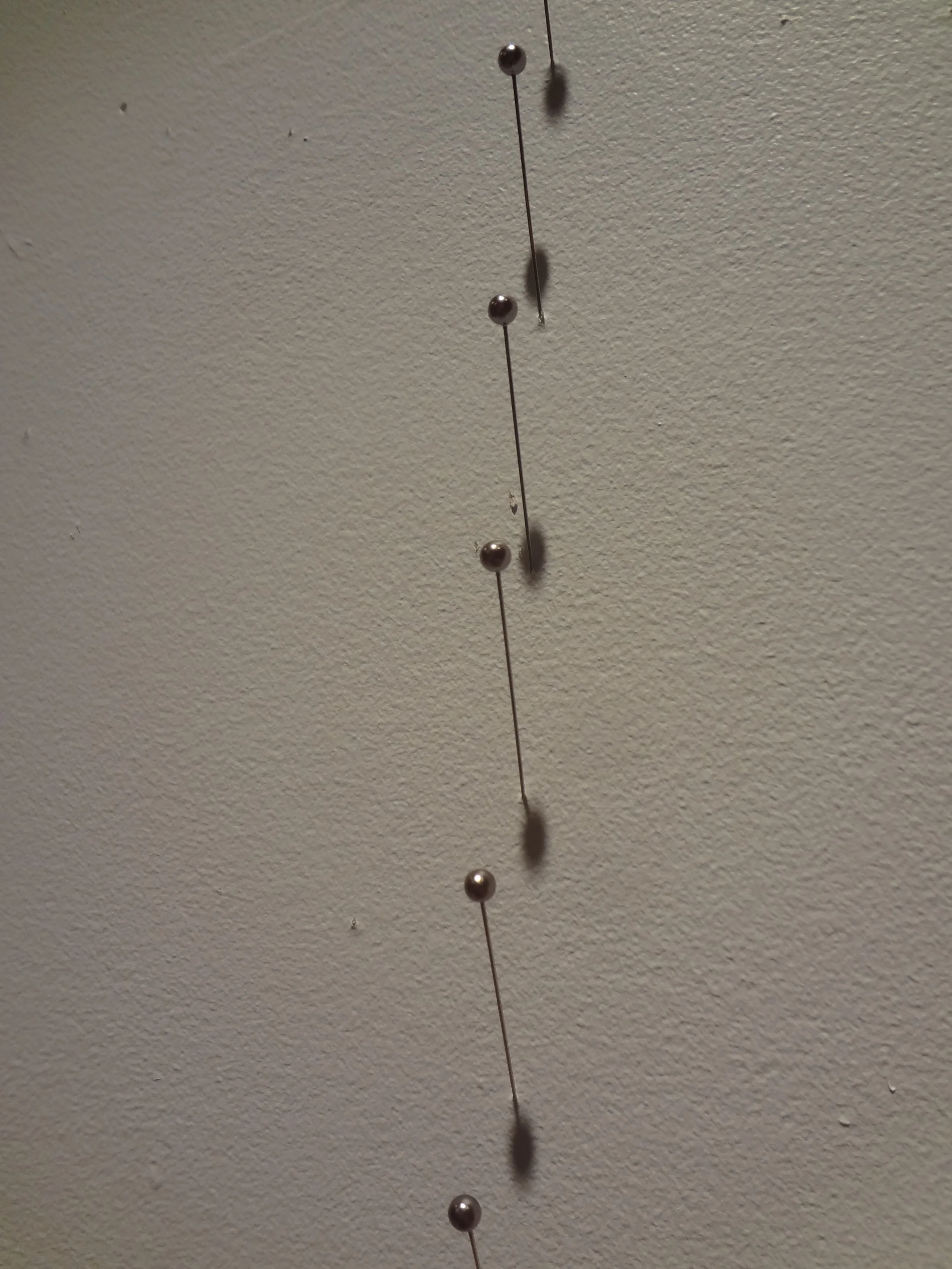 A vertical string with metallic pins, each casting shadows on a beige wall.