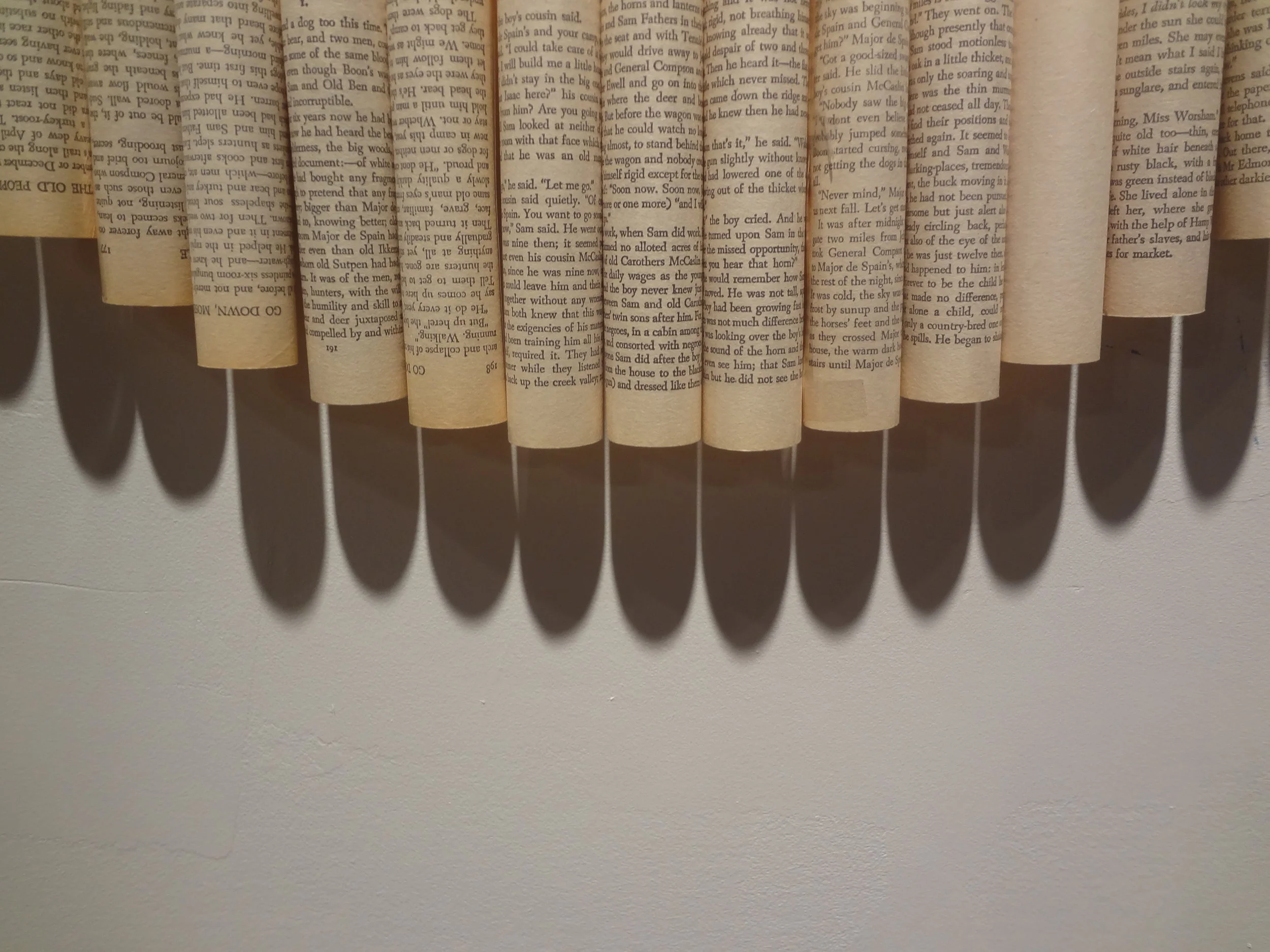 An art piece made from rolled pages of a book arranged in a circular pattern, casting shadows on a white wall.