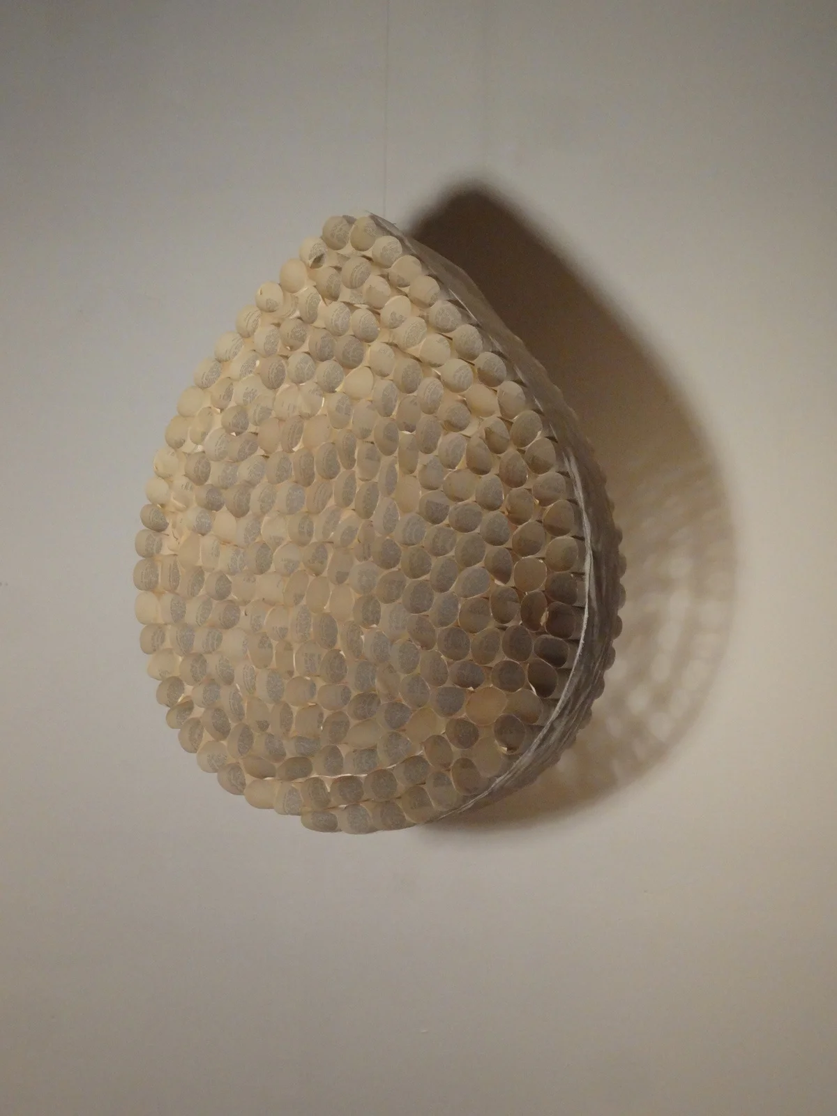 A circular wall art piece made of what appears to be numerous small, cream-colored paper tubes arranged in a dome shape, casting a shadow on a plain light-colored wall.