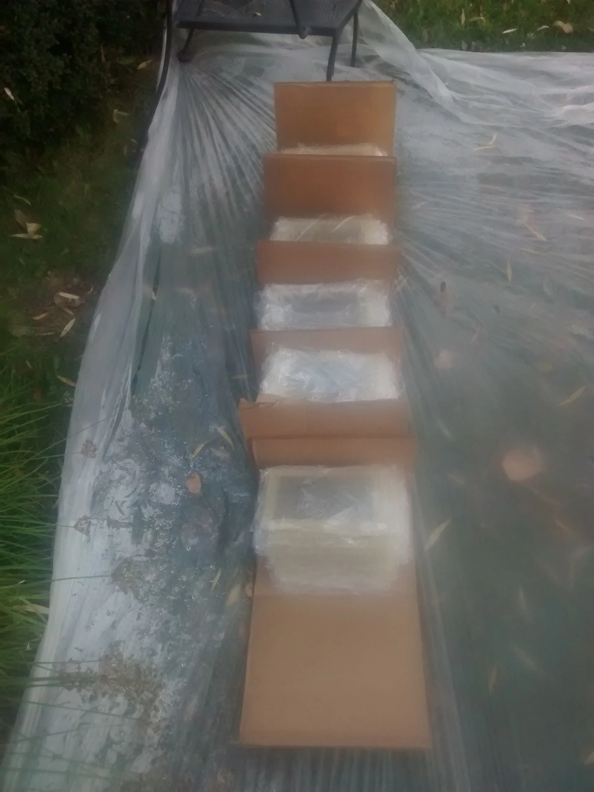 Row of partially unwrapped bricks on a black plastic sheet outdoors, with plastic covering nearby and greenery in the background.