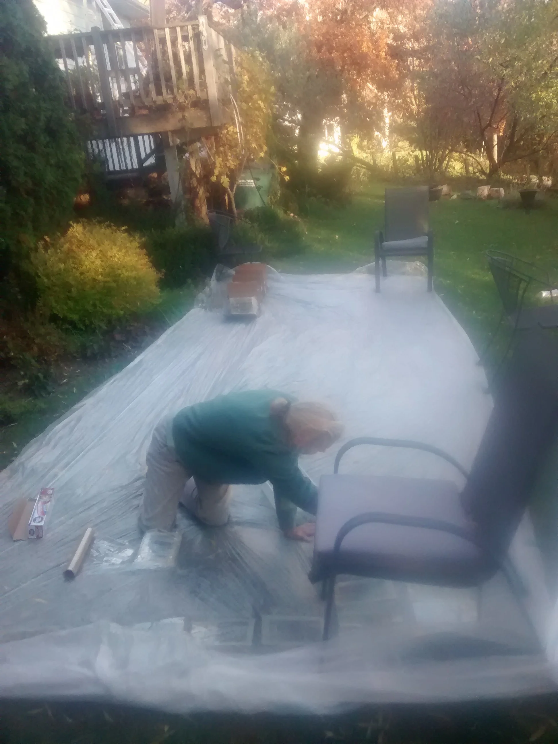 A man is kneeling on a plastic-covered outdoor patio during daytime, working on a chair with a paint roller nearby. There are patio furniture items and trees with autumn foliage in the background.