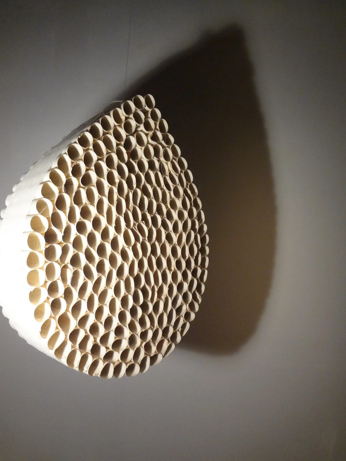 A light-colored circular wooden sculpture with numerous small cylindrical tubes arranged in a pattern, casting a shadow on the wall.