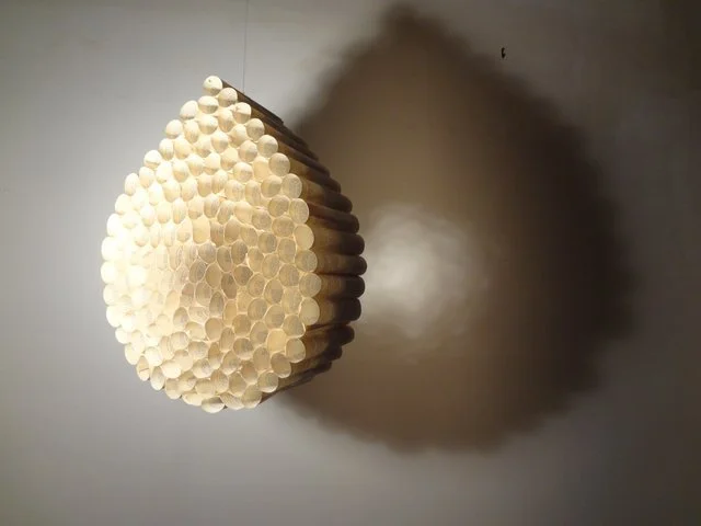 A wall sculpture made of vertically arranged wooden sticks creating a flattened, three-dimensional shape resembling a honeycomb or abstract form, casting a shadow on a gray wall.