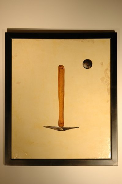 Abstract art piece resembling a face with a vertical wooden element as a nose, a small dark circle above it as an eye, and a dark horizontal element at the bottom as a mouth, framed in black on a beige background.
