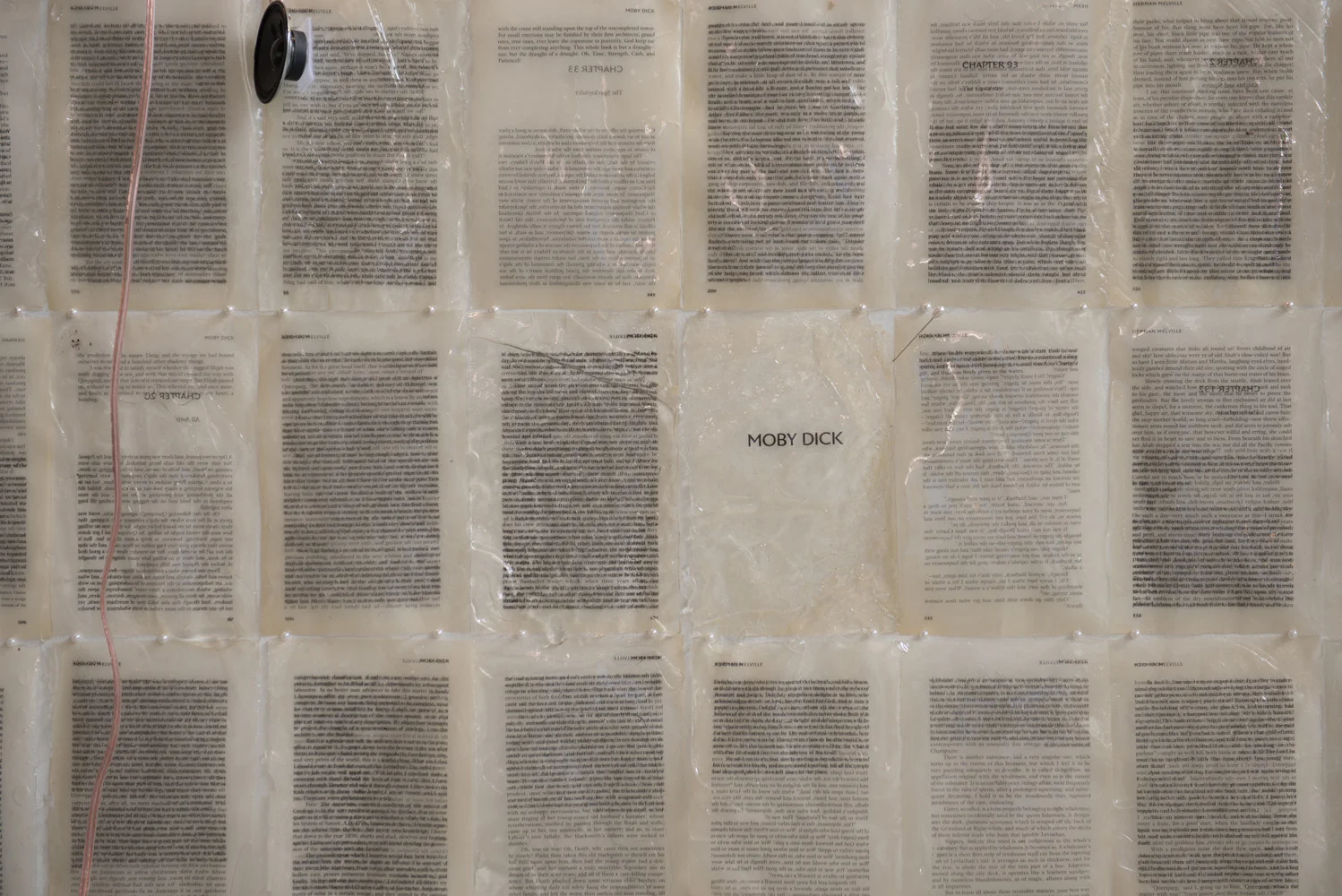 A wall covered with multiple pages of text in plastic sleeves, with a central page labeled "MOBY DICK." In the upper left corner, there's a small speaker with a cable hanging downward.