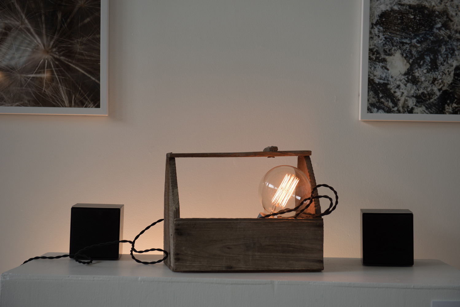 Decorative lamp with Edison bulb in a wooden box, flanked by black speakers on a white surface, with abstract art pictures on a white wall in the background.