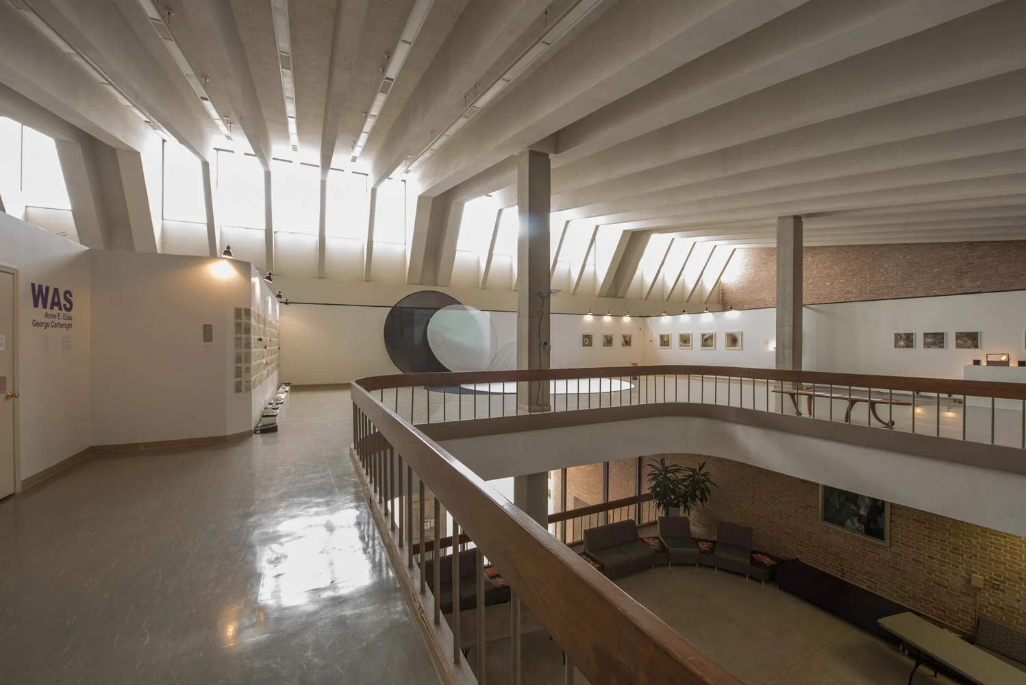 Interior of an art gallery with curved railing, artwork on walls, a large round sculpture, and skylights illuminating the space.