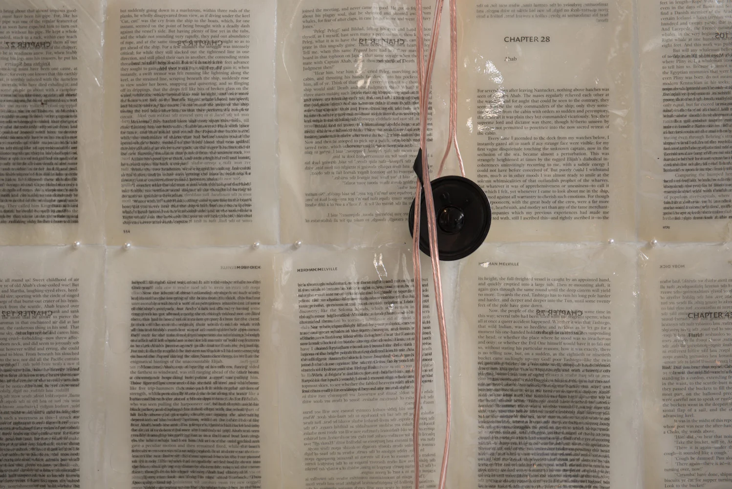A black speaker with pink and white wires is mounted on a transparent plastic sheet covering several pages of text from a book or magazine, laid flat on a surface.
