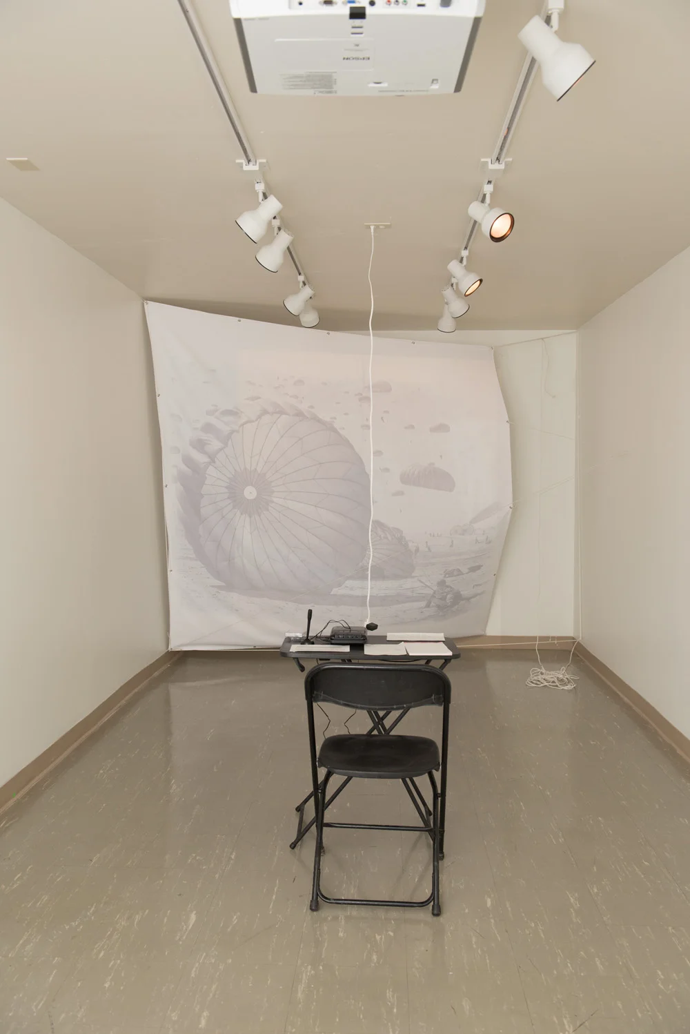 Empty art studio with a small black folding chair, a table with electronic equipment, and a large photograph of a parachute projected on a white backdrop. Track lighting illuminates the space, and a ceiling-mounted projector is visible.