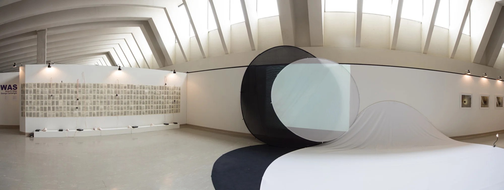 Modern art installation in a spacious gallery, featuring a black cylinder with an attached curved white structure, and artwork on the walls