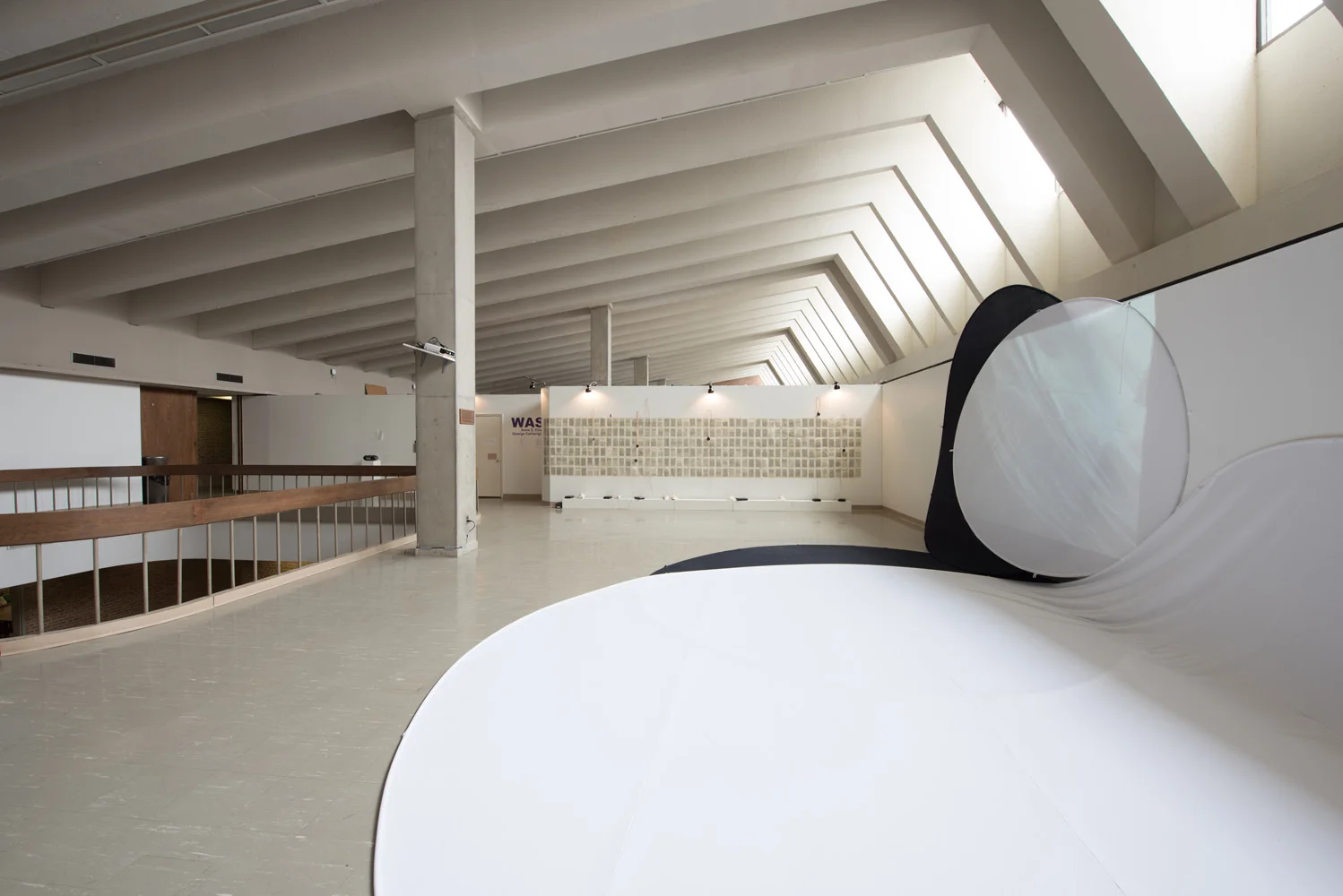 Empty modern interior space with a curved white installation or sculpture in the foreground, a wall with small square displays in the background, and a large sloped ceiling with exposed beams.