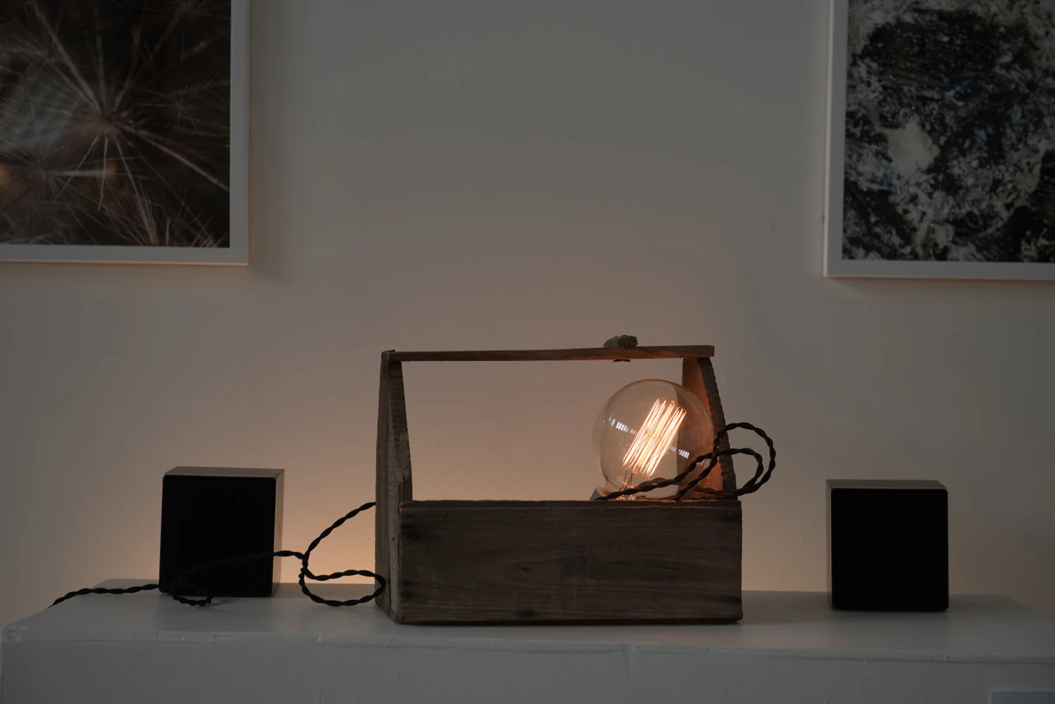 Decorative light bulb with filament inside a wooden box, with black speakers on either side, on a white surface against a plain wall, with framed artwork hanging above.